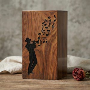 May include: A wooden box with a carved silhouette of a musician playing a trumpet, with musical notes emanating from the instrument. The box is made of a dark wood with a visible grain pattern. The design is a decorative item.