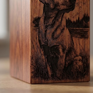 Puede incluir: Una urna rectangular de madera con una ilustraci&oacute;n grabada con l&aacute;ser de un perro sentado en hierba alta cerca de un lago con un bosque al fondo. La veta de la madera es visible y el grabado muestra texturas detalladas de pelaje y paisaje.
