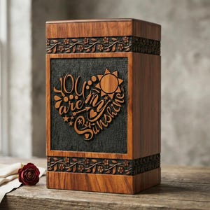 Puede incluir: Una caja de madera con intrincados grabados florales y las palabras "You are my sunshine" en un diseño en forma de corazón. La caja tiene un acabado de madera oscura con un panel negro contrastante en el centro.