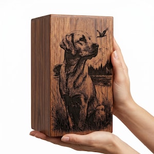 Puede incluir: Una urna rectangular de madera con una ilustraci&oacute;n grabada a l&aacute;ser en tono oscuro de un Labrador sentado en hierba alta. El fondo muestra un lago, un bosque de pinos y un ave volando frente a un paisaje monta&ntilde;oso.