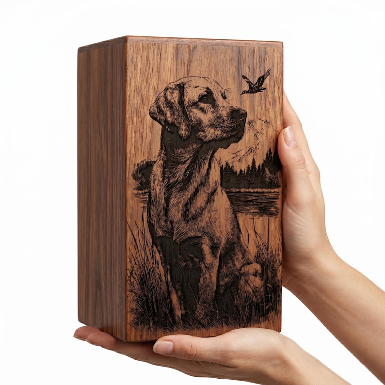 Puede incluir: Una urna rectangular de madera con una ilustraci&oacute;n grabada a l&aacute;ser en tono oscuro de un Labrador sentado en hierba alta. El fondo muestra un lago, un bosque de pinos y un ave volando frente a un paisaje monta&ntilde;oso.