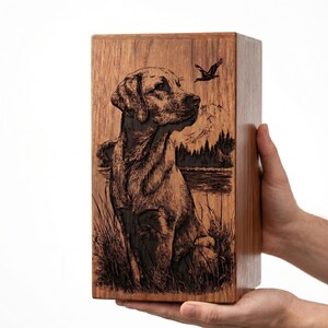 Puede incluir: Una urna rectangular de madera con una ilustraci&oacute;n grabada con l&aacute;ser de un Labrador sentado en hierba alta. La escena incluye un bosque, un cuerpo de agua y un ave en vuelo sobre un fondo de monta&ntilde;as, todo en tonos de color marr&oacute;n oscuro.
