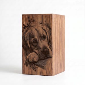 May include: A rectangular wooden urn featuring a detailed laser-engraved portrait of a golden retriever dog resting its head on its paws. The natural wood grain is visible across the surface, with dark brown etched lines defining the dog's features.