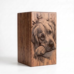 May include: A rectangular wooden urn featuring a detailed, dark brown laser-engraved portrait of a golden retriever dog resting its head on its paws. The natural wood grain is visible across the surface, showcasing light and dark brown tones.