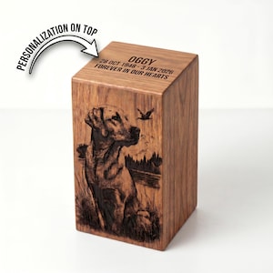 Puede incluir: Una urna rectangular de madera con una imagen grabada a l&aacute;ser de un Labrador en un paisaje con un p&aacute;jaro. La superficie superior tiene grabado el texto OGGY, 28 OCT 1948 - 3 JAN 2026 y FOREVER IN OUR HEARTS, con una flecha personalization on top.