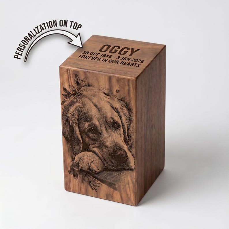 May include: A rectangular wooden urn featuring a laser-engraved portrait of a dog on the front panel. The top surface is engraved with the name OGGY, the dates 28 OCT 1948 - 3 JAN 2026, and the text FOREVER IN OUR HEARTS. A graphic arrow points to the top.