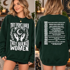 Könnte beinhalten: Ein dunkelgrünes Langarm-Sweatshirt mit weißen Grafikdrucken. Die Vorderseite zeigt Hände, die eine Mondsichel halten, mit dem Text "THEY DIDN'T BURN WITCHES THEY BURNED WOMEN." Die Rückseite listet historische Daten und Meilensteine für Frauen auf.