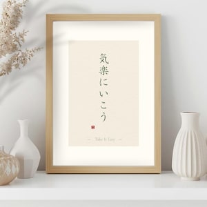 以下が含まれることがあります： 日本語の文字と英語のフレーズ「Take It Easy」が書かれた額入りのプリント。アートワークは、濃い緑色のテキストが入った明るいベージュの背景が特徴です。フレームは明るい木の色で、プリントは装飾的な花瓶のある白い棚に飾られています。