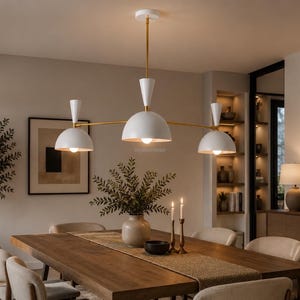 May include: A three-light chandelier featuring white dome-shaped shades with gold-toned accents and a central gold-toned rod. The fixture is suspended from a white ceiling canopy and provides downward illumination over a dining area.