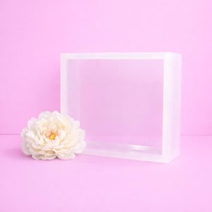 Könnte beinhalten: Eine transparente, quadratische Acryl-Ausstellungsbox auf einer rosa Oberfläche, mit einer weißen Pfingstrose links. Die Box hat ein minimalistisches Design, ideal für die Präsentation von Produkten oder Dekoration.