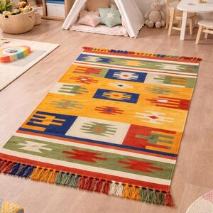 May include: A rectangular, colorful area rug with a yellow background and geometric patterns in red, blue, green, and white. The rug has a fringe of tassels in various colors along the short edges. This rug is suitable for a child's room.