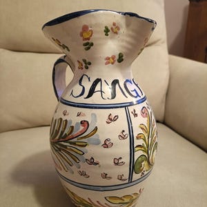 May include: White ceramic jug with a blue rim and handle, decorated with colourful floral and leaf patterns. The word "SANG" is written in blue script. A decorative and functional piece for serving drinks.