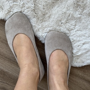 Puede incluir: Un par de bailarinas de ante gris claro. Los zapatos tienen una puntera redondeada y un perfil bajo. Las bailarinas se muestran sobre una alfombra clara y texturizada y un suelo de madera.