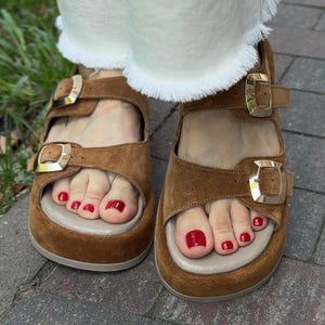 May include: Brown suede sandals with gold buckles. The sandals have a beige footbed and are worn with white pants with a frayed hem. The toenails are painted red.