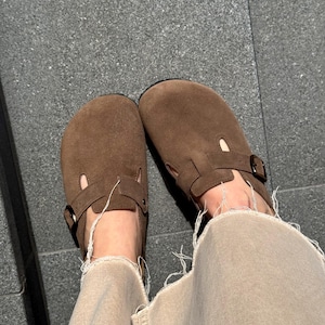 May include: A pair of brown suede clogs with adjustable buckles. The clogs have a rounded toe and a strap across the top. The image shows the clogs from a top-down perspective, with the wearer's legs visible.