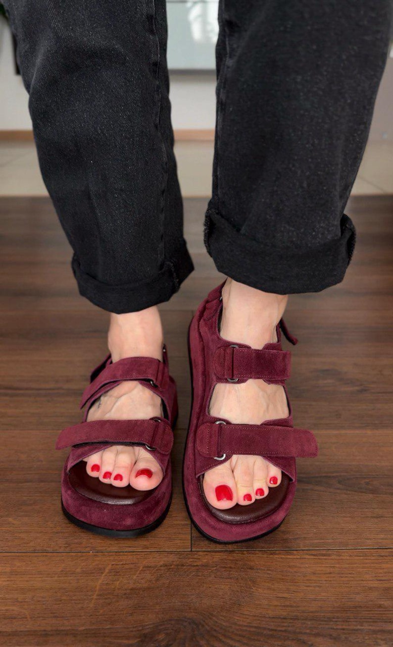 May include: Burgundy suede sandals with adjustable straps. The sandals have a contoured footbed and are worn with black denim jeans. The image shows a close-up of the feet and sandals.