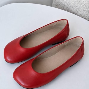 May include: A pair of bright red flat shoes with a rounded toe design and a smooth finish. The interior of the shoes is lined with a light beige material, and they feature a low-profile silhouette with a thin black sole visible at the heel.