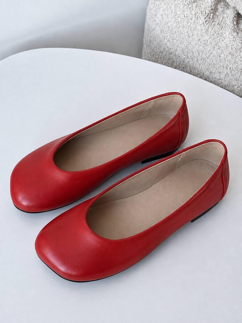 May include: A pair of bright red flat shoes with a rounded toe design and a smooth finish. The interior of the shoes is lined with a light beige material, and they feature a low-profile silhouette with a thin black sole visible at the heel.