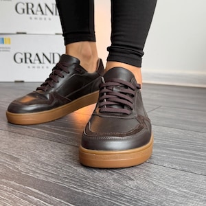 Puede incluir: Zapatillas de cuero marrón con cordones marrones y suela color canela. Las zapatillas las lleva una persona con leggings negros. El fondo incluye cajas con el texto "GRAND SHOES".