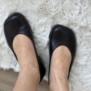 Puede incluir: Un par de bailarinas de cuero negro con punta cuadrada. Los zapatos se usan sobre una alfombra clara y texturizada. Las bailarinas tienen un perfil bajo y un diseño sencillo, adecuado para el uso diario.