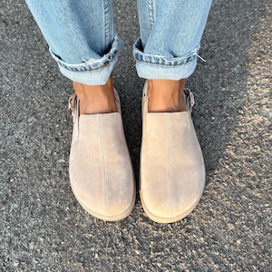 Può includere: Un paio di scarpe slip-on in camoscio beige con dettagli fibbia. Le scarpe sono indossate con jeans di denim blu chiaro con risvolti. Le scarpe hanno una punta arrotondata e un tacco basso.