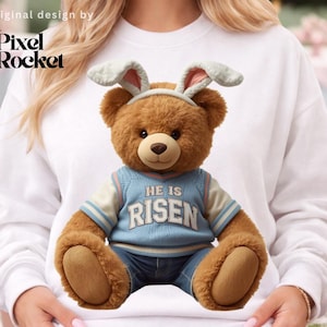 May include: A brown teddy bear wearing a blue vest with the text "HE IS RISEN" and denim jeans. The bear has a headband with bunny ears. The Pixel Rocket logo is in the upper left corner. The bear is sitting on a white sweatshirt.