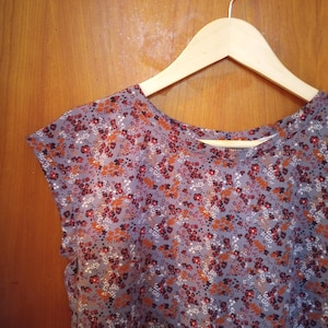 May include: A cap-sleeved top with a floral pattern in shades of gray, red, orange, and white hangs on a wooden hanger. The fabric is covered in small, detailed flower designs, creating a delicate and feminine aesthetic. The top is made of a lightweight material.