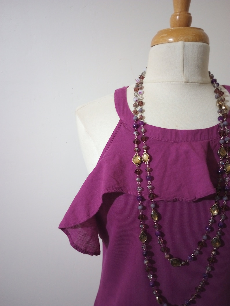 May include: A magenta sleeveless top with a ruffled neckline and a layered beaded necklace with purple, brown, and gold accents.