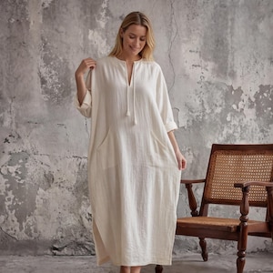 Puede incluir: Un vestido midi de color blanco roto con mangas largas, escote en V con cierre de lazo central, dos bolsillos laterales y una silueta holgada y fluida. La prenda está confeccionada en tejido texturizado y llega a media pantorrilla con aberturas.
