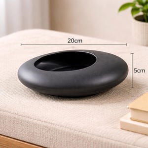 May include: A black ceramic bowl, oval in shape, measuring 20cm long and 5cm high. The bowl has a smooth, rounded exterior and a deep, open interior. It is displayed on a neutral-toned surface, suitable for decorative or practical use.