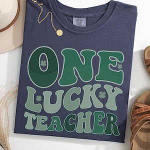 Navy blue Comfort Colors 1717 t-shirt with "One Lucky Teacher" green graphic print. Distressed vintage style teacher shirt for St. Patrick’s Day celebrations, shown in a stylish flat lay.