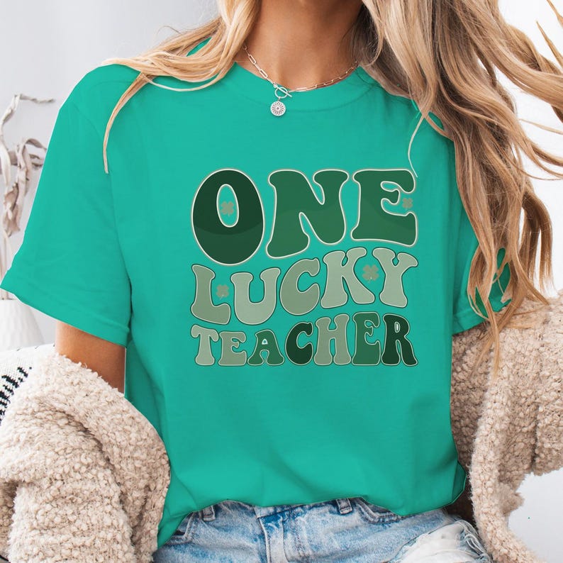 Female model wearing a İsland green Comfort Colors 1717 t-shirt with "One Lucky Teacher" retro print. Styled with a cozy cardigan and jeans for a festive St. Patrick's Day classroom outfit.