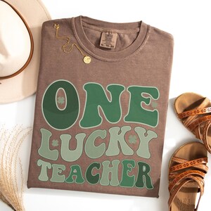 Comfort Colors 1717 Espresso t-shirt featuring a retro "One Lucky Teacher" design in shades of green with shamrocks. Flat lay styling with a boho hat and sandals, perfect St. Patrick's Day teacher gift idea.