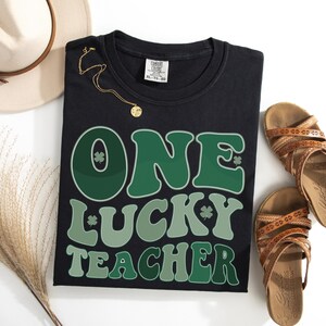 Black Comfort Colors 1717 garment-dyed shirt with a "One Lucky Teacher" shamrock design. Retro aesthetic teacher appreciation gift, minimalist flat lay photography.