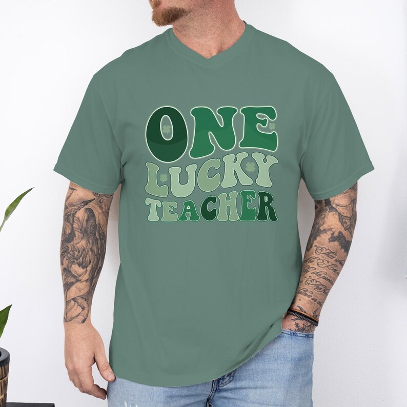 Male model with tattoos wearing a Comfort Colors 1717 Light Green t-shirt. Retro "One Lucky Teacher" design, oversized fit, casual St. Patrick's Day apparel for male teachers.