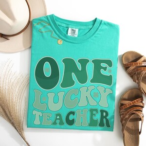 İsland Green Comfort Colors 1717 shirt with vintage "One Lucky Teacher" wavy typography. St. Paddy's Day outfit for teachers, styled in a flat lay with summer accessories for a trendy classroom look.