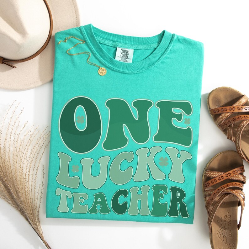 İsland Green Comfort Colors 1717 shirt with vintage "One Lucky Teacher" wavy typography. St. Paddy's Day outfit for teachers, styled in a flat lay with summer accessories for a trendy classroom look.