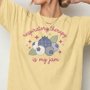 May include: A pale yellow t-shirt featuring a graphic of blueberries with various patterns including plaid, gingham, and polka dots. The text reads respiratory therapy is my jam in a pink script font, surrounded by small pink and green star accents.
