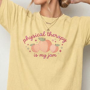 Könnte beinhalten: Ein blassgelbes Kurzarm-T-Shirt mit einer Grafik aus drei Pfirsichen im Vichy-Karo-Muster. Der Text "physical therapy is my jam" ist in einer rosa Schreibschrift gedruckt, umgeben von kleinen Sternsymbolen und grünen Blättern.