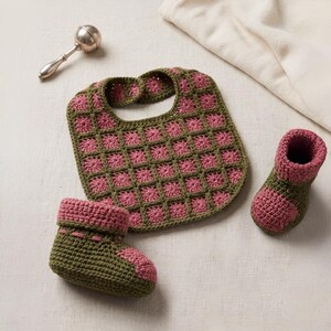 Può includere: Un set per neonati all'uncinetto fatto a mano in verde oliva e rosa, composto da un bavaglino e due scarpine. Il bavaglino e le scarpine presentano un motivo quadrato. È visibile anche un sonaglio d'argento.