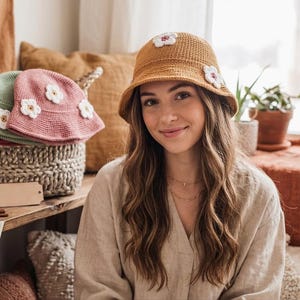 Peut inclure: Une femme porte un chapeau de pêcheur au crochet jaune moutarde orné d'applications de fleurs blanches et roses. Le chapeau fait partie d'une collection de chapeaux au crochet faits à la main, comprenant des chapeaux roses et verts, exposés dans un panier tressé.