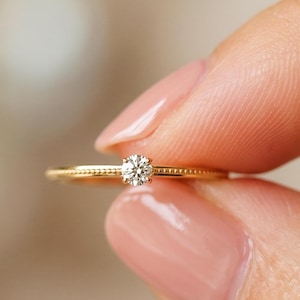 Puede incluir: Un anillo fino de oro con un pequeño diamante de talla brillante redonda engastado en el centro. La banda presenta una delicada textura de cuentas a lo largo de su superficie, lo que aporta un aspecto refinado y detallado al metal de esta joya.