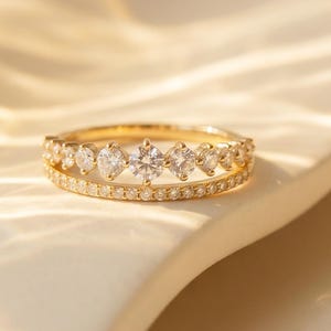 May include: A gold ring featuring two bands joined together. The top band is set with a series of round and pear-shaped clear stones, while the bottom band is a thin, delicate pavé-style band encrusted with tiny, sparkling clear stones.