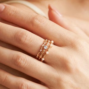 May include: Three gold-colored rings stacked on a finger. The rings feature delicate designs with small pearls and a central light blue gemstone. The rings are worn on a hand with manicured nails.