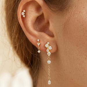 May include: Three gold-toned earrings featuring clear, sparkling stones. One earring is a small cluster stud, another is a delicate drop stud, and the third is a longer dangling earring with a chain and a pear-shaped stone at the bottom.