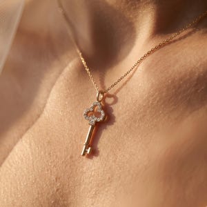 May include: A gold-toned necklace featuring a pendant shaped like a skeleton key. The bow of the key is designed in a clover-like shape and is encrusted with small, clear, sparkling stones. The chain is a delicate, thin gold-toned metal link.