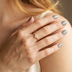 Può includere: Un anello d'oro con un motivo floreale di piccole pietre preziose rotonde e trasparenti. L'anello è su un dito, con la mano e il braccio visibili. La fascia dell'anello è sottile e delicata.