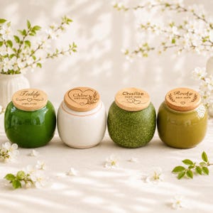 May include: Four small, round ceramic urns with wooden lids, each engraved with a name and dates. The first is glossy green, the second is white, the third is speckled green, and the fourth is olive green. Each lid features a heart motif.