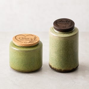 Pode incluir: Dois potes cilíndricos de cerâmica com esmalte verde sálvia manchado. O pote mais baixo tem uma tampa de madeira clara gravada com um coração, ramos de folhas e o texto Chloe 2012-2024. O pote mais alto tem uma tampa de madeira escura com Rocky 2017-2025.