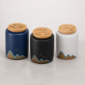 May include: Three cylindrical ceramic urns with wooden lids. The blue, black, and white urns feature mountain and bird graphics at the base. The lids are engraved with names Bailey, Daisy, and Chloe, along with dates and decorative motifs.